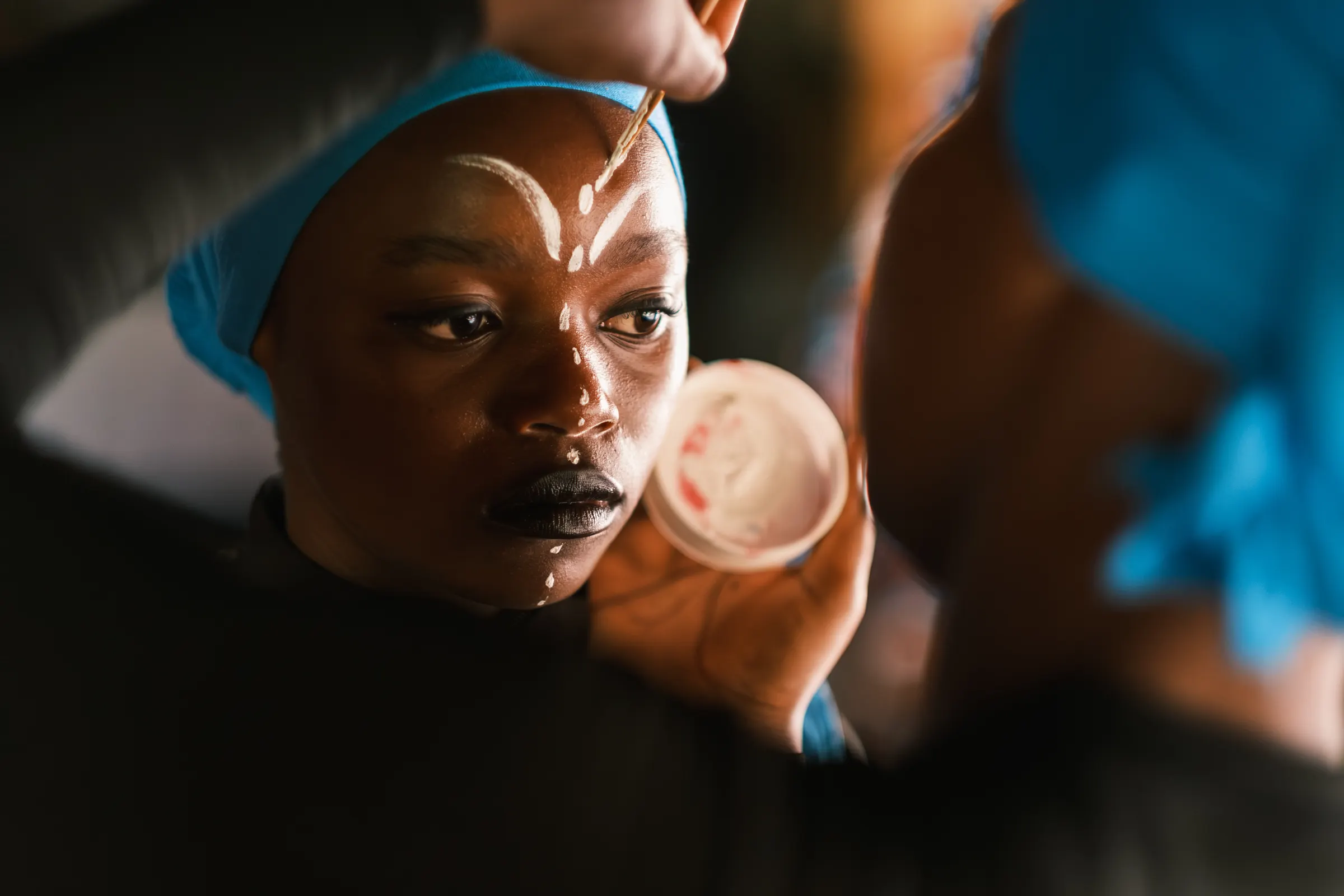 A performer sitting still while white markings are painted across her brow before a performance