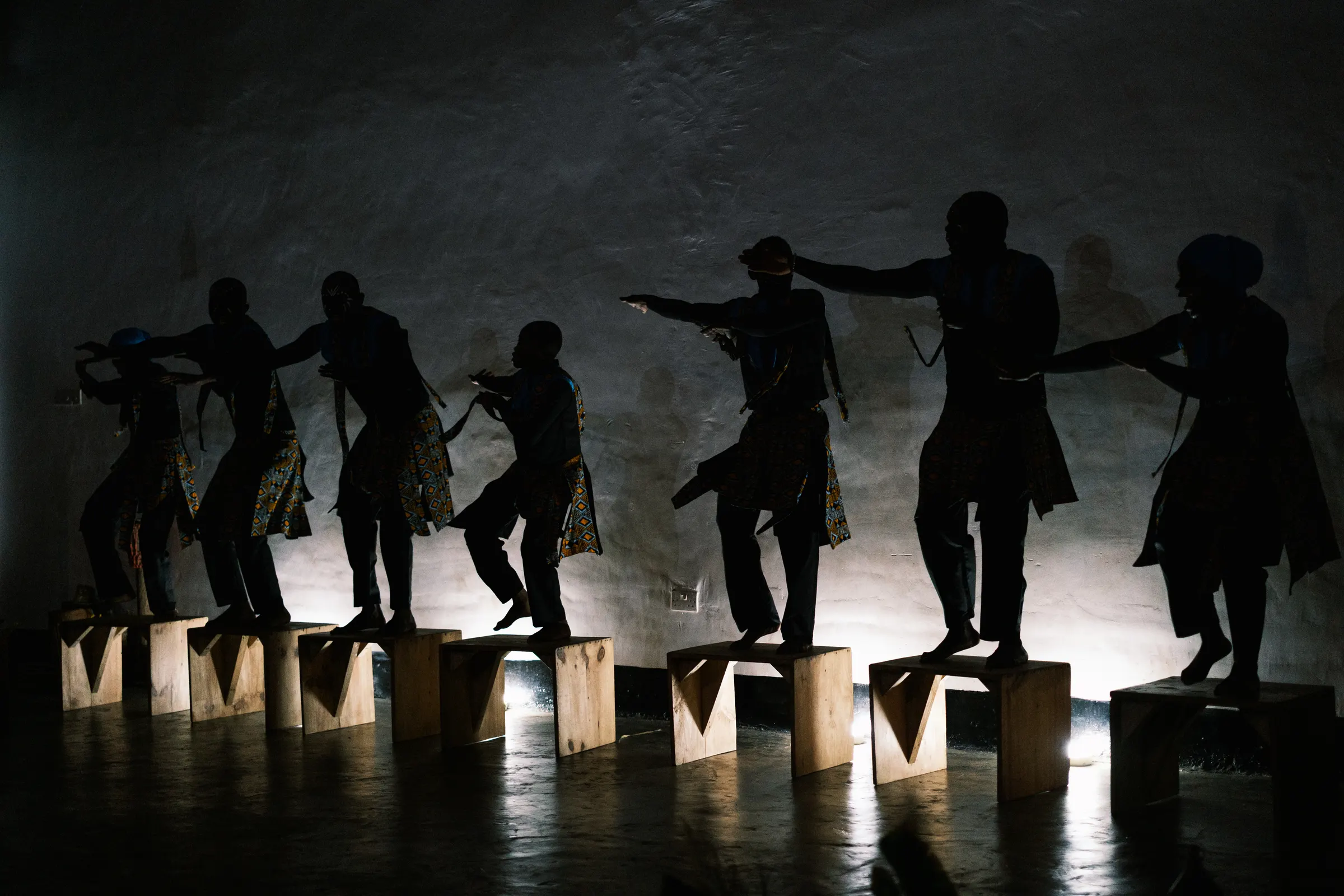 Dancers in silhouette, arms outstretched, balanced on a line of wooden plinths against a lit wall