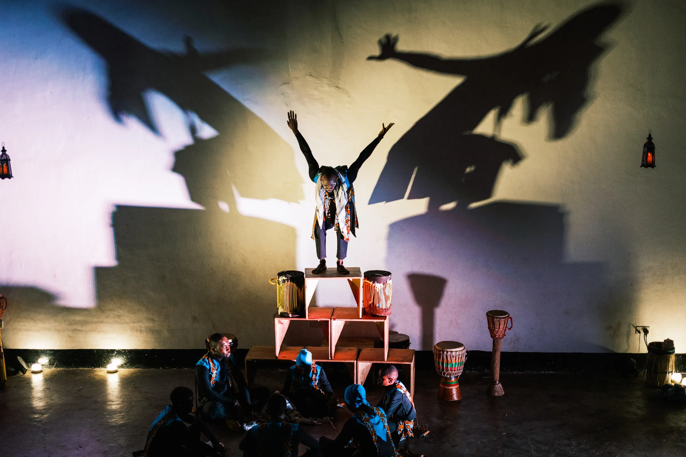 A performer standing on a stack of drums with arms raised, casting large shadows across the wall while the ensemble watches from the floor